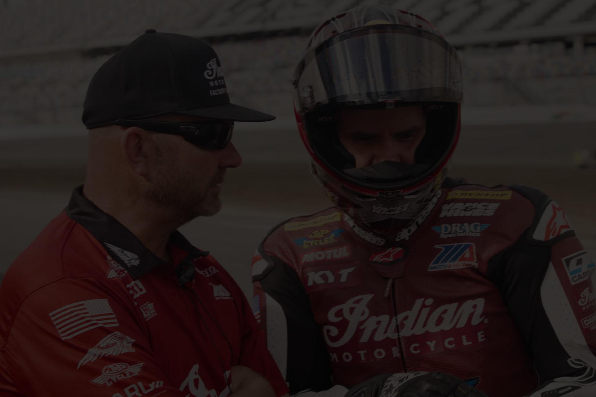 Two men in red motorcycle racing gear talk in the pit lane; one wears a helmet and visor.](https-//)